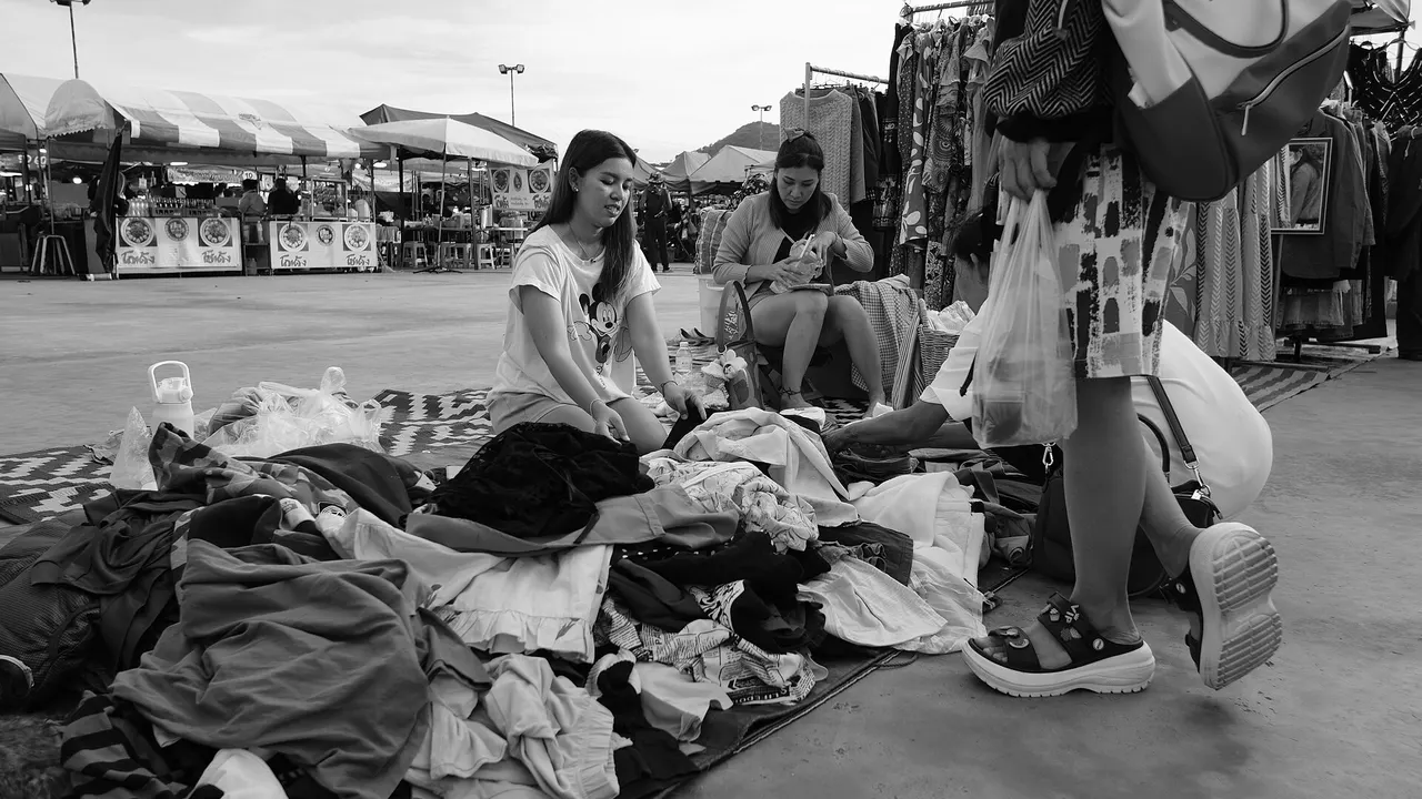 Shoppers sifting through a large pile of secondhand clothes on the ground at an open-air market, with stalls and umbrellas behind